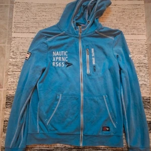 Blå hoodie med dragkedja Nautic XPRNC - Säljer en blå hoodie från Nautic XPRNC med hel dragkedja, huva och tryck på bröstet och ärmen. Tröjan har tre fickor med dragkedja och sportig look. Perfekt för dig som gillar en avslappnad stil.