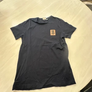 Svarta Burberry t-shirts med logga - Två stilrena svarta t-shirts från Burberry med klassisk rund hals och kort ärm. Ena t-shirten har en beige patch med Burberrys monogram på bröstet, den andra har en svart broderad logga. Båda är i mjuk bomull och passar perfekt till jeans eller shorts.
