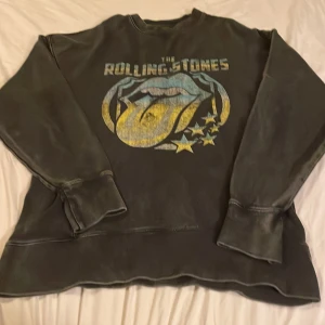 Svart Rolling Stones sweatshirt Pull&Bear - Cool svart sweatshirt från Pull&Bear med stort Rolling Stones-tryck i gult och blått framtill. Tröjan har lång ärm, rund hals och mjuk fleece på insidan. Perfekt för dig som gillar bandmerch och vill ha en avslappnad stil.