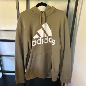 Beige hoodie från Adidas med logga - Snygg beige hoodie från Adidas med stor vit logga framtill. Tröjan har huva med snörning, känguruficka och långa ärmar. Perfekt för en avslappnad och sportig stil.
