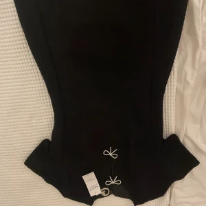 Svart kort klänning med rosetter - Supersnygg svart kort klänning från Next med figurnära passform och coola detaljer som glittrande rosetter framtill. Klänningen har en unik form vid nederkanten och är gjord i återvunnen polyester. Perfekt för dig som vill sticka ut med stil. Aldrig använt, köpt för: ca 500kr