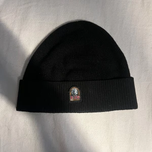 Svart ribbstickad mössa Parajumpers - Svart ribbstickad mössa från Parajumpers med uppvikt kant och klassisk PJS-logga framtill. Baksidan har en röd etikett med Parajumpers-tryck. Perfekt för kalla dagar och snygg till streetwear.