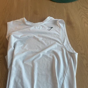 Vit Gymshark linnetopp i S - Säljer en vit linnetopp från Gymshark i storlek S. Toppen är ärmlös och har en sportig design med diskret Gymshark-logga på bröstet. Materialet är lätt och andas, perfekt för träning eller gym. Snygg och enkel att matcha med dina favoritträningskläder.