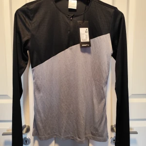 Svart och grå långärmad träningströja Craft - Sportig långärmad tröja från Craft i svart och ljusgrått. Tröjan har rund hals, diagonal färgdelning och är tillverkad i ett snabbtorkande, stretchigt syntetmaterial som andas. Perfekt för träning och aktiva dagar.