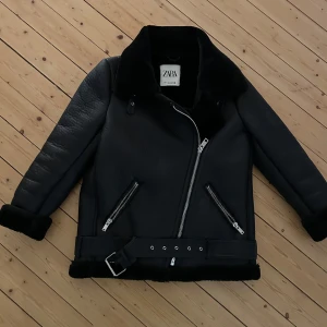 Svart bikerjacka från Zara med fuskpäls på insidan - Säljer en svart bikerjacka från Zara i storlek S. Jackan har en schysst  krage, dragkedja i silver, två sneda fickor med dragkedjor och ett brett bälte nertill. Insidan är fodrad med mjuk fuskpäls. 