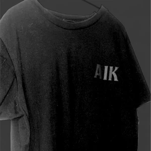 Svart AIK t-shirt med tryck - Svart t-shirt från AIK med stort klubbmärke tryckt på framsidan och AIK-logga på bröstet. Klassisk rund hals och korta ärmar. Perfekt för dig som vill visa ditt stöd för AIK. Materialet är mjukt och skönt, troligtvis bomull. Inga hål och den är inte solblekt den är fin 
