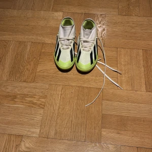 Adidas vita och limegröna gympaskor  - Säljer ett par Adidas fotbollsskor i vitt och limegrönt med svarta ränder på sidorna. Skorna har snörning och låg profil, perfekt för inomhusspel. Materialet är syntet och designen är sportig och modern.