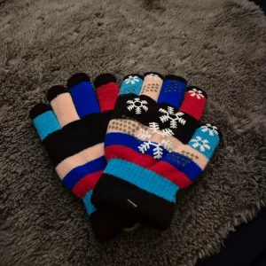 Färgglada stickade vantar med snöflingor - Säljer ett par stickade vantar i flera färger med randigt mönster och vita snöflingor på ovansidan. Vantarna har inslag av svart, blått, rött, beige och turkost. Perfekta för kalla vinterdagar och ger en lekfull touch till din outfit.