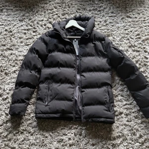 Svart pufferjacka från Jd sport märket är supply and demand  - Svart pufferjacka från supply and demand  med huva och dragkedja framtill. Jackan har två sidofickor med dragkedja och quiltad design. Perfekt för kalla dagar och har en clean, stilren look som funkar till allt.