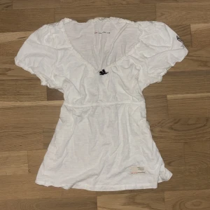 Vit blus med v-ringning från Odd Molly - Supersöt vit blus från Odd Molly med v-ringning, kort puffärm och liten rosett framtill. Blusen har broderad logga på ena ärmen och volangkant vid hals och ärmslut. Perfekt för en fräsch och avslappnad look.