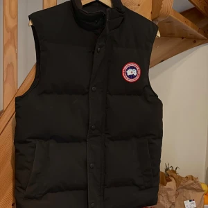 Svart dunväst från Canada Goose - Svart dunväst från Canada Goose med klassisk logotyp på bröstet. Västen har en quiltad design, två sidofickor och knäppning framtill. 