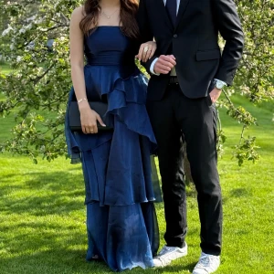 Balklänning med volanger - Säljer en marinblå balklänning med flera lager volanger och axelbandslös design. Klänningen har en snygg snörning i ryggen och är tillverkad i ett glansigt, lätt material som ger en lyxig känsla. Perfekt för bal! Fråga gärna om fler bilder eller mått. Klänningen är lite trasig längst ner men inget som syns mycket, och går lätt att laga med bara en enkel söm! På lappen står det storlek L men den passar mig perfekt som vanligtvis brukar ha S. Klänningen är bara använd en gång på min egna bal!🤗