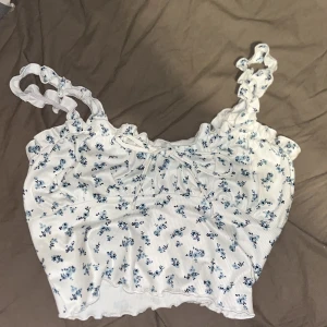 Vit blommig linnetopp från SHEIN - Supersöt vit linnetopp från SHEIN med små blå blommor, volangkant och rynkade axelband. Toppen har en söt knytning framtill och är croppad med vågig nederkant. Perfekt för en somrig look!