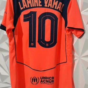 Barcelona Lamine Yamal orange matchtröja - Säljer en Barcelona fotbollströja med Lamine Yamal och nummer 10 på ryggen. Tröjan är orange med mörkblå detaljer, Nike-logga, Spotify och UNHCR tryck. Kortärmad modell i lätt och ventilerande material, perfekt för match eller träning.