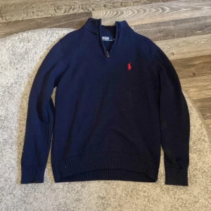 Marinblå stickad tröja Polo Ralph Lauren - Snygg marinblå stickad tröja från Polo Ralph Lauren med röd broderad logga på bröstet. Tröjan har half zip, ribbade muddar och hög krage. Perfekt för dig som gillar klassisk och stilren look. Nypris 1599kr