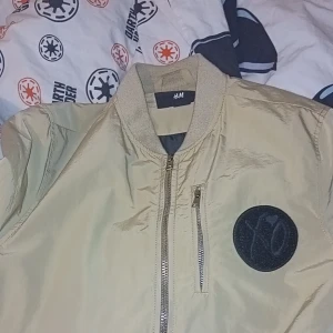 Beige bomberjacka XO H&M - Snygg beige bomberjacka från H&M med svart XO-tryck på ryggen och en rund svart patch framtill. Jackan har dragkedja och ribbade muddar vid krage och ärmslut. Perfekt för dig som vill sticka ut med en streetstyle-look.