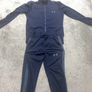 Marinblå Under Armour set - Snygg marinblå träningsväst från Under Armour med dragkedja framtill och diskret logga på bröstet. Västen har en sportig look och är gjord i lätt syntetmaterial som andas. Perfekt för gymmet eller löprundan.