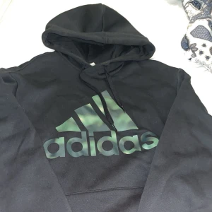Svart Adidas hoodie med logga - Svart hoodie från Adidas med stor logga i grönt och svart framtill. Klassisk huva med snörning och mjukt material som känns skönt mot huden. Perfekt för dig som gillar sportig och avslappnad stil. S men passar även M