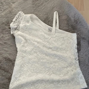 Vit spetsig oneshoulder topp - Säljer en vit topp med spets och blommigt mönster. Toppen har en axelbandslös design med en smal axelrem på ena sidan och är oneshoulder. Perfekt för dig som gillar romantiska detaljer och vill sticka ut lite extra.