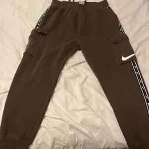 Bruna cargopants från Nike - Bruna mjukisbyxor från Nike och vita små och stora Nike detaljer. Byxorna har svarta och vita band längs benen med små Nike-loggor. Tillverkade i mjukt material och har resår i midjan och benslut. En av dom 4 knaparna som finns på fickorna går inte att knäppa