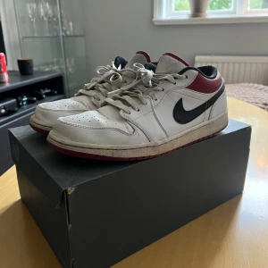 Nike Air Jordan 1 Low vit/röd/svart - Nike Air Jordan 1 Low sneakers i vitt skinn med svarta swoosh-loggor och röda detaljer runt hälen. Skorna har röd yttersula, svart foder och klassisk snörning. 