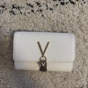 Vit axelväska från Valentino - Snygg vit axelväska från Valentino med guldfärgad kedja och stort V i metall på framsidan. Väskan har en stilren, rektangulär form och är gjord i skinn med lätt textur. Dekorativ tofs hänger under loggan för extra detalj. Perfekt för dig som gillar lyxiga accessoarer. Observera att själva ”kedjorna” som hänger under märket har tappat lite av sin färg.
