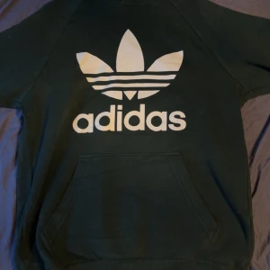 Svart Adidas hoodie med stor logga - Säljer en svart hoodie från Adidas med den klassiska stora vita trefoil-loggan och text på bröstet. Tröjan har huva med snörning och en stor magficka. Perfekt för dig som gillar streetwear och sportig stil.