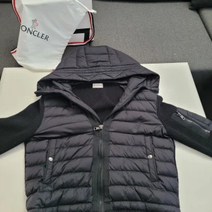Svart moncler cardigan  - Svart cardigan från Moncler med stickade ärmar och huva. Västen har quiltad framsida, två fickor med tryckknappar och dragkedja framtill. Moncler-logga på ärmen och extra ficka med dragkedja på ena ärmen. Perfekt för dig som gillar exklusiv streetstyle.