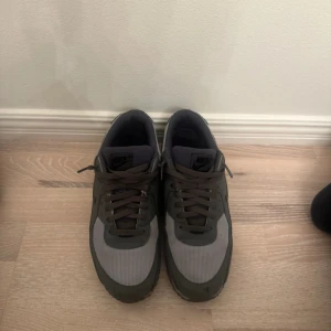 Nike Air Max 90 i grå och svart - Snygga Nike Air Max 90 sneakers i olika nyanser av grått och svart. Ovandelen är i mesh och syntet med svarta detaljer och klassisk Air Max-sula. Skorna har snörning och en platt, mönstrad sula för bra grepp. Perfekta för dig som gillar stilrena och sportiga sneakers.