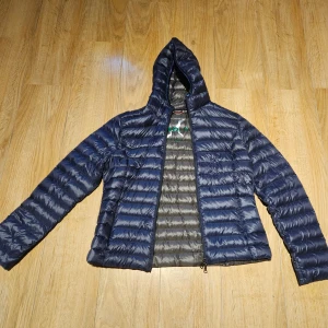 Mörkblå dunjacka , no 1  - Säljer en mörkblå, quiltad dunjacka  strl L , med huva och dragkedja framtill. PASSAR ALLA ÅRSTIDER . Jackan har en slim passform och är perfekt för kyliga dagar. Märkeslogga på ärmen och snygg, stilren look som funkar till det mesta. För trång över bysten för mig. Köpt I nyskick från sellpy, aldrig använt själv . 