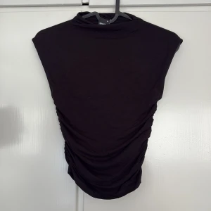 Svart topp från Gina Tricot - En svart topp från Gina Tricot i storlek XS. Toppen har en enkel design med rynkade sidor och är ärmlös, vilket ger en snygg och modern siluett. Perfekt för dig som gillar minimalistisk stil och vill ha något som sitter snyggt.