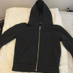 Svart hoodie med dragkedja XS - Klassisk svart hoodie i storlek XS med hel dragkedja framtill och stor huva. Tröjan har två rymliga fickor på magen och ribbade muddar vid ärmslut och nederkant. Perfekt för en chill och avslappnad stil.