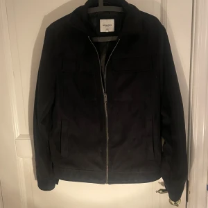 Svart jacka från Jack & Jones - Stilren svart jacka från Jack & Jones med dragkedja framtill och två bröstfickor med lock. Jackan har en klassisk krage och raka ärmar, perfekt för dig som gillar en clean look. Materialet känns slitstarkt och passar till många outfits.