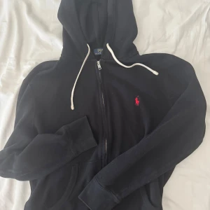 Svart hoodie från Polo Ralph Lauren - Svart hoodie från Polo Ralph Lauren med dragkedja, vit snörning i huvan och klassisk röd broderad logga på bröstet. Mjuk bomullskänsla och känguruficka framtill. Perfekt för en chill och stilren look.