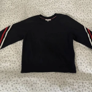 Mörkblå Tommy Hilfiger tröja med ränder - Snygg mörkblå tröja från Tommy Hilfiger med röda och vita ränder längs ärmarna och över bröstet. Klassisk crewneck och diskret logga på ena ärmen. Perfekt till jeans eller kjol för en sportig vibe.