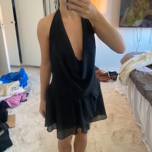 Svart kort klänning med öppen rygg - Supersnygg svart kort klänning med djup urringning och öppen rygg som knyts i nacken. Klänningen har volangkant nertill och är tillverkad i ett lätt, flowy material som ger en cool vibe. Perfekt för dig som vill sticka ut med en stilren och trendig look.