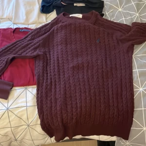 Vinröd kabelstickad tröja från Peak One - Säljer en vinröd kabelstickad tröja från Peak One med rund hals och diskret blå logga broderad på bröstet. Tröjan har lång ärm och ribbade muddar, perfekt för dig som gillar klassisk och stilren look.