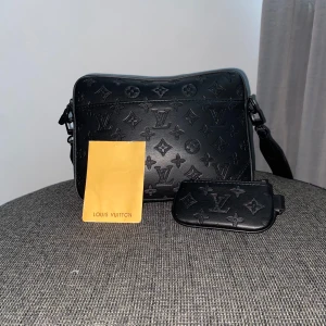 Svart axelväska  - Säljer en svart axelväska i skinn med klassiskt monogrammönster. Väskan har justerbar axelrem och medföljande liten plånbok. Perfekt för dig som vill ha en stilren och lyxig accessoar.