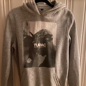 Grå hoodie med Tupac-tryck - Snygg grå hoodie från H&M med stort svartvitt Tupac-tryck på bröstet. Tröjan har huva, känguruficka och långa ärmar. Perfekt för dig som gillar streetstyle och vill ha en bekväm och stilren look. Den har hål i fickan!!