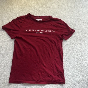 Vinröd t-shirt från Tommy Hilfiger - Snygg vinröd t-shirt från Tommy Hilfiger med klassisk logga och text framtill. T-shirten har rund halsringning och korta ärmar. Tillverkad i mjuk bomull som är skön att ha på sig. Perfekt för en chill och stilren look. Kommer strykas innan skickning!