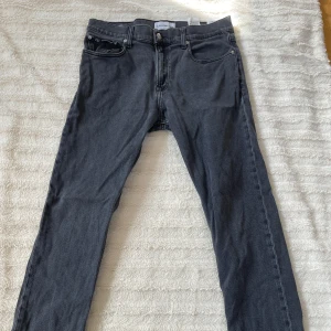 Mörkgrå Calvin Klein jeans - Snygga mörkgrå jeans från Calvin Klein med klassisk femficksdesign och CK-logga på bakfickan. Modellen har raka ben och normal passform, tillverkade i bomull med lite stretch för extra komfort. Perfekt för dig som gillar stilrena och tidlösa jeans.