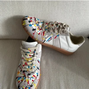 Maison Margiela sneakers med färgstänk - Unika sneakers från Maison Margiela med vit bas och beige detaljer i mocka. Skorna har coola färgstänk i rött, blått, gult och svart över hela ovandelen. (pris kan diskuteras vid snabb affär) ALLT MED FÖLJER SOM SKOLÅDA O DUSTBAG✅