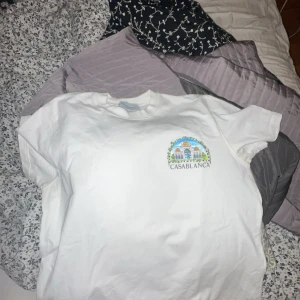 Vit Casablanca t-shirt med tryck - Vit t-shirt från Casablanca med färgglatt tryck av byggnader och palmer på ryggen samt mindre logga på bröstet. Klassisk passform och rund hals. Perfekt statement-plagg för dig som gillar unika prints och streetstyle. Passar m L