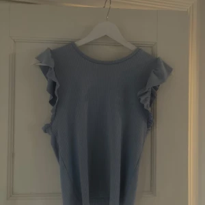 Ljusblå ribbad topp från Stradivarius - Supersöt ljusblå ribbad topp från Stradivarius med volangdetaljer på axlarna. Toppen är ärmlös och har en rund halsringning. På sidan är något slags hål med för mycket stygn. det kan nog lösas med att ta bort stygnen!