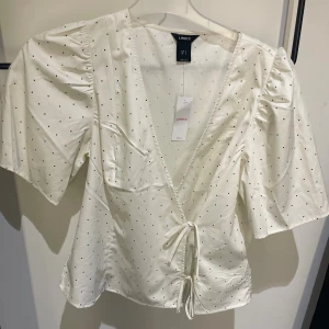 Vit prickig omlottblus från Lindex - Supersöt vit omlottblus från Lindex med svarta prickar och knyt framtill. Blusen har v-ringning, korta puffärmar och lätt glansigt material som känns lyxigt. Perfekt för dig som gillar en feminin och trendig look.