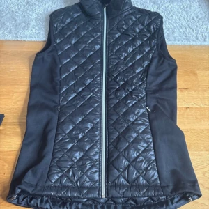 Svart väst konståkning  - Snygg svart dunväst från JIV Sport i storlek S. Västen har ett quiltat rutmönster framtill och baktill, dragkedja hela vägen och mjuka sidopaneler i fleece. Perfekt för dig som gillar sportig och stilren look. Den är avsedd till att användas vid träning vid konståkning men kan även användas i vardag eller andra sporter! Aldrig använd men saknar prislapp!