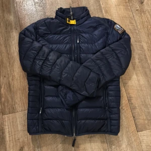 Mörkblå dunjacka från Parajumpers M - Säljer en mörkblå dunjacka från Parajumpers i storlek M. Jackan är superlätt, har quiltad design, hög krage med gul detalj och dragkedja framtill. Klassisk patch på ärmen och flera praktiska fickor. Perfekt för kyliga dagar när du vill hålla stilen.