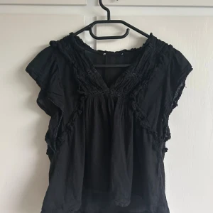 Svart blus med volangdetaljer - Svart blus med v-ringning och volangdetaljer längs axlar och bröst. Lös passform och korta ärmar ger en avslappnad vibe. Perfekt för dig som gillar en bohemisk och cool stil.