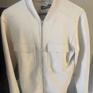 Vit cardigan från Dressmann - Snygg vit/beige cardigan från Dressmann i storlek S. Jackan har två stora fickor med lock framtill men även innerfickor, ribbad krage och muddar samt dragkedja hela vägen. Materialet känns mjukt och har en clean look som passar till många stilar. Nypris 799. Mitt pris 499. 