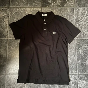 Svart pikétröja från Lacoste - Klassisk svart pikétröja från Lacoste med korta ärmar och krage. Tröjan har knäppning med två knappar framtill och den ikoniska broderade krokodilloggan på bröstet. Tillverkad i bomull för en skön och luftig känsla. Priset kan diskuteras. Helt ny  